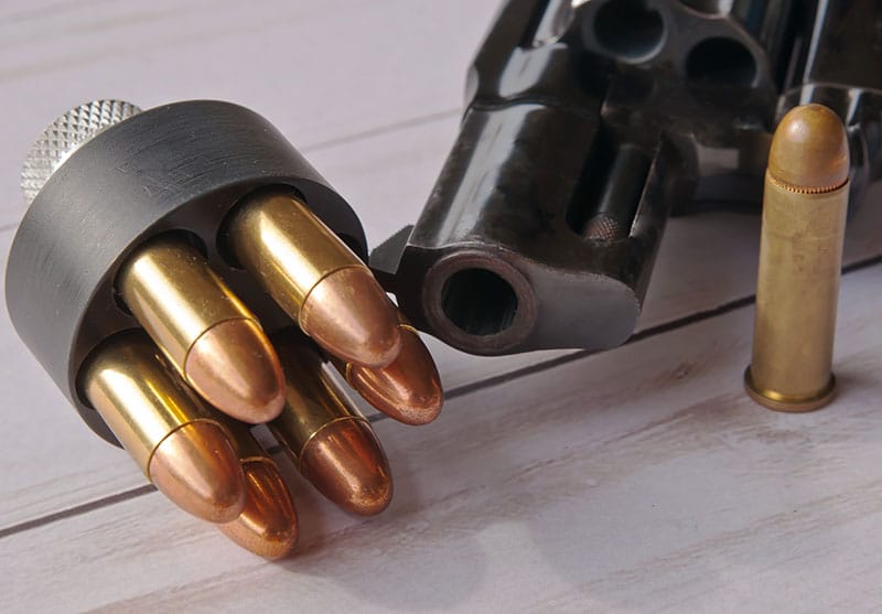 A loaded speed loader loaded with 38 special full metal jacket bullets in front of a black revolver with a single bullet